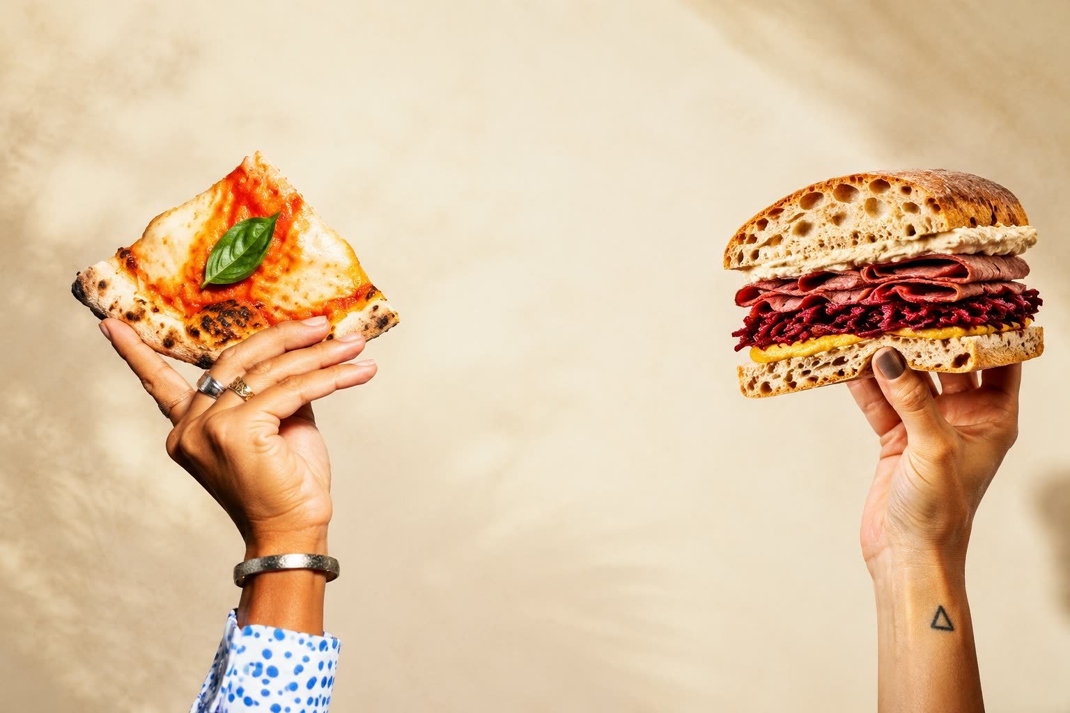 Mâini ridicând o felie de pizza și un sandwich cu pastrami GIO, pe fundal crem